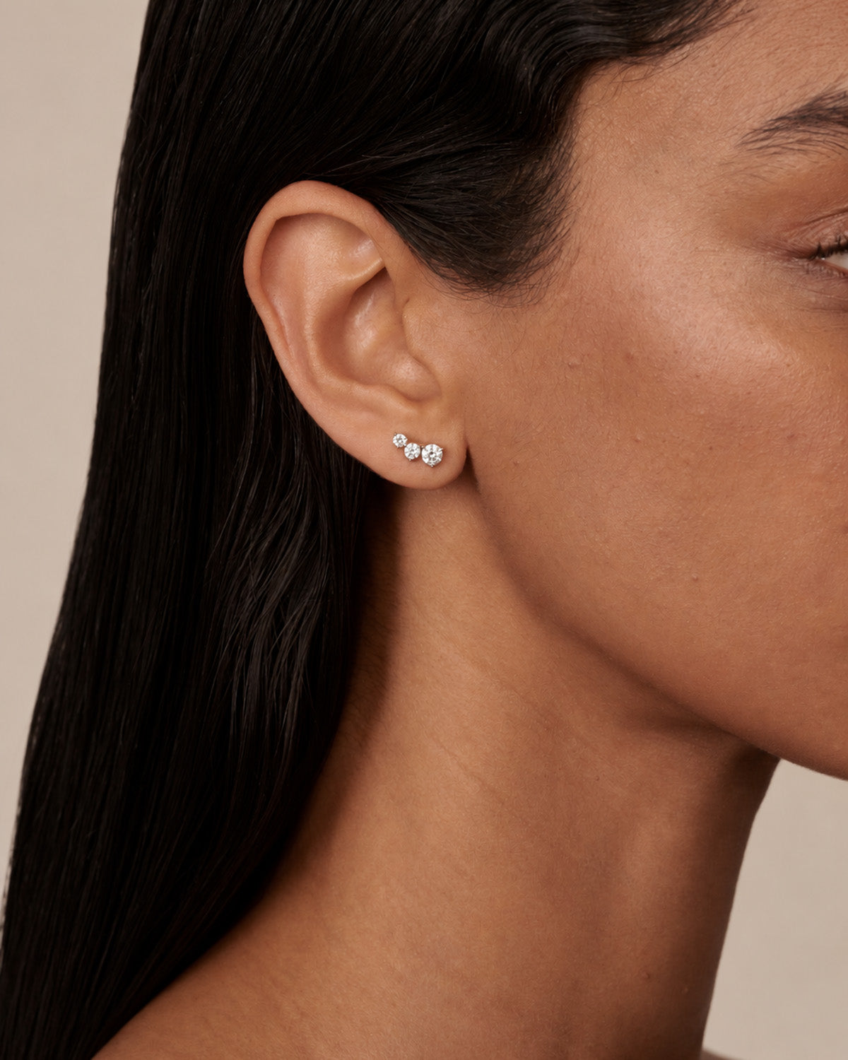 A close-up of a model's ear wearing a small silver stud earring with three clear diamonds, set against a neutral beige background.