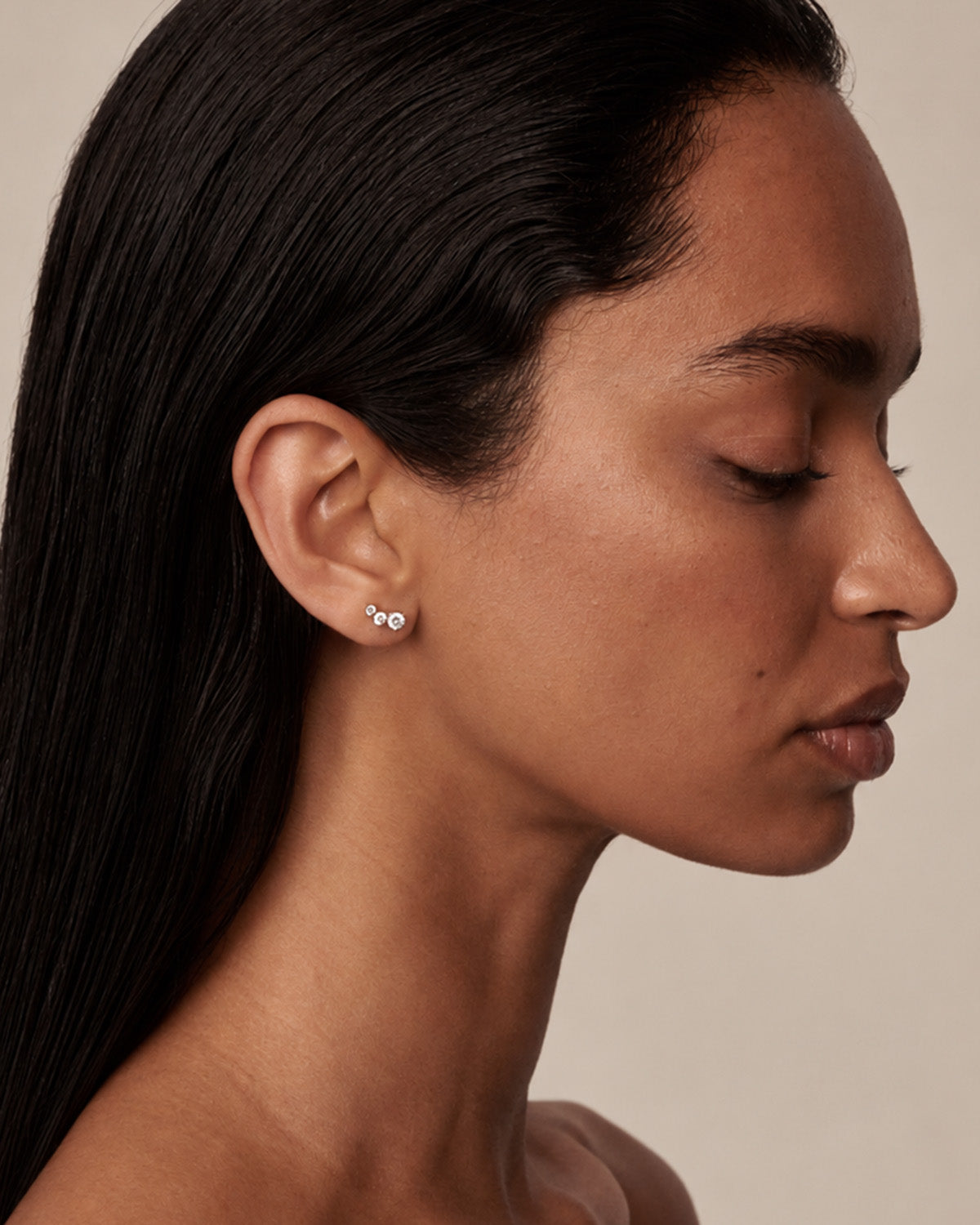 A close-up side profile of a woman with dark hair wearing a small cluster stud earring with three clear diamonds in a silver setting, against a warm beige background.