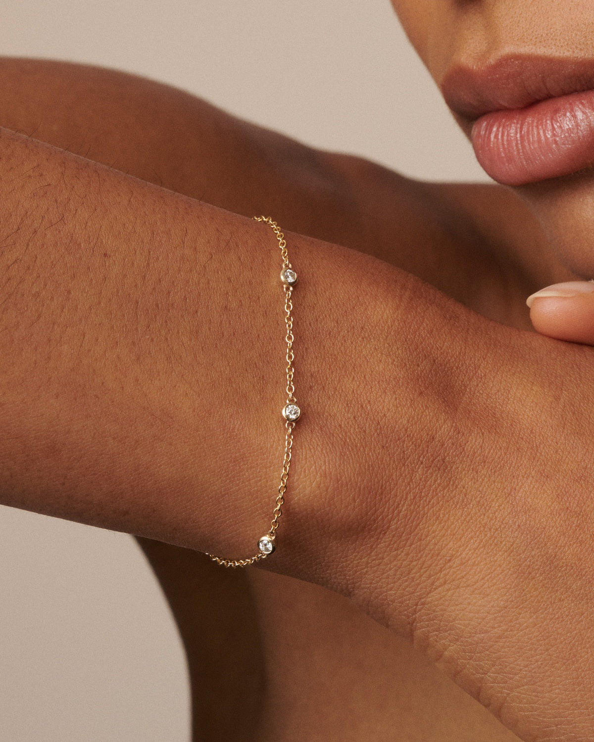 A delicate gold chain bracelet with three small clear crystal beads, shown worn on a person's wrist against a warm beige background.