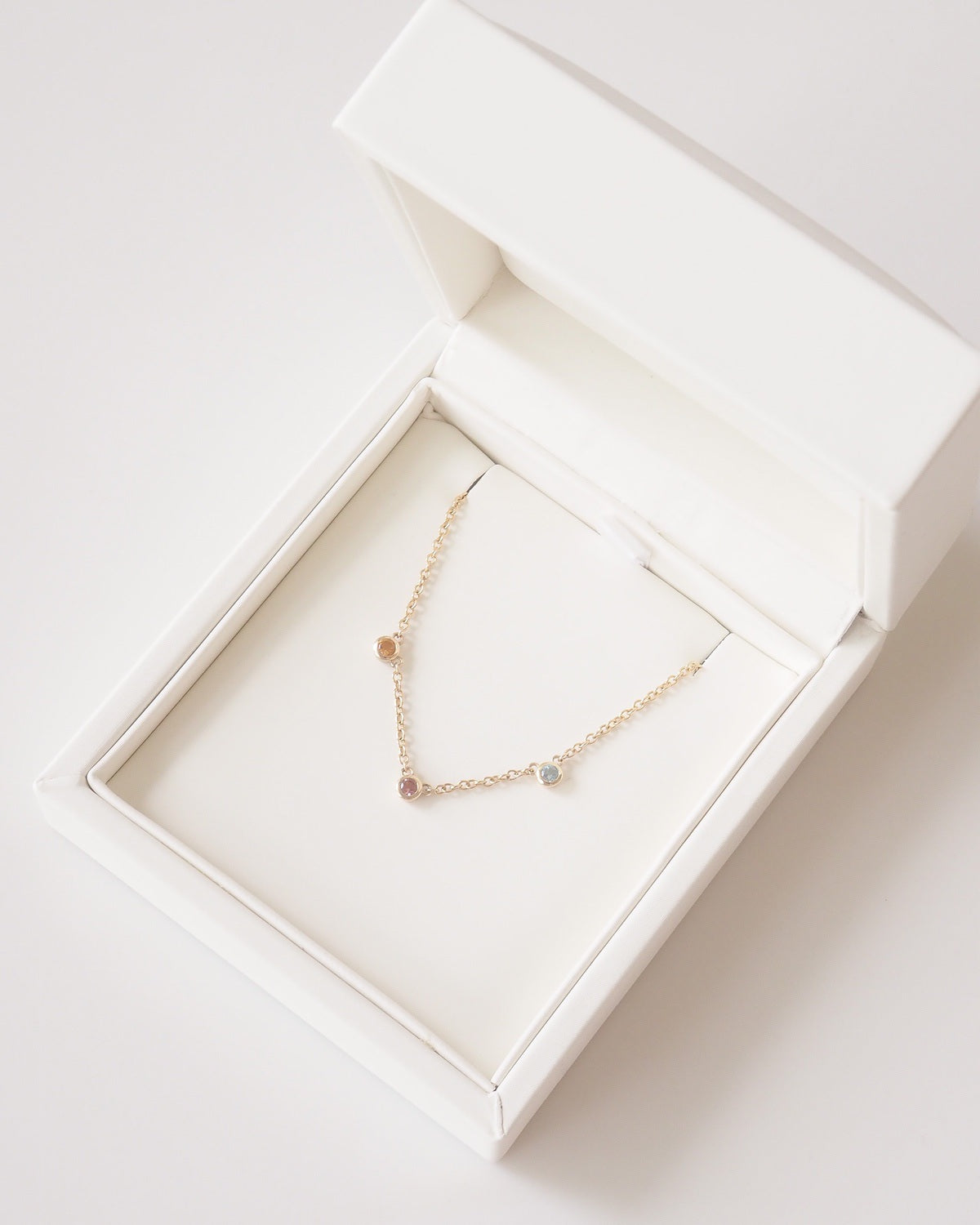 A delicate gold chain necklace with small, multi-colored gemstone charms, presented inside an open white jewelry box.