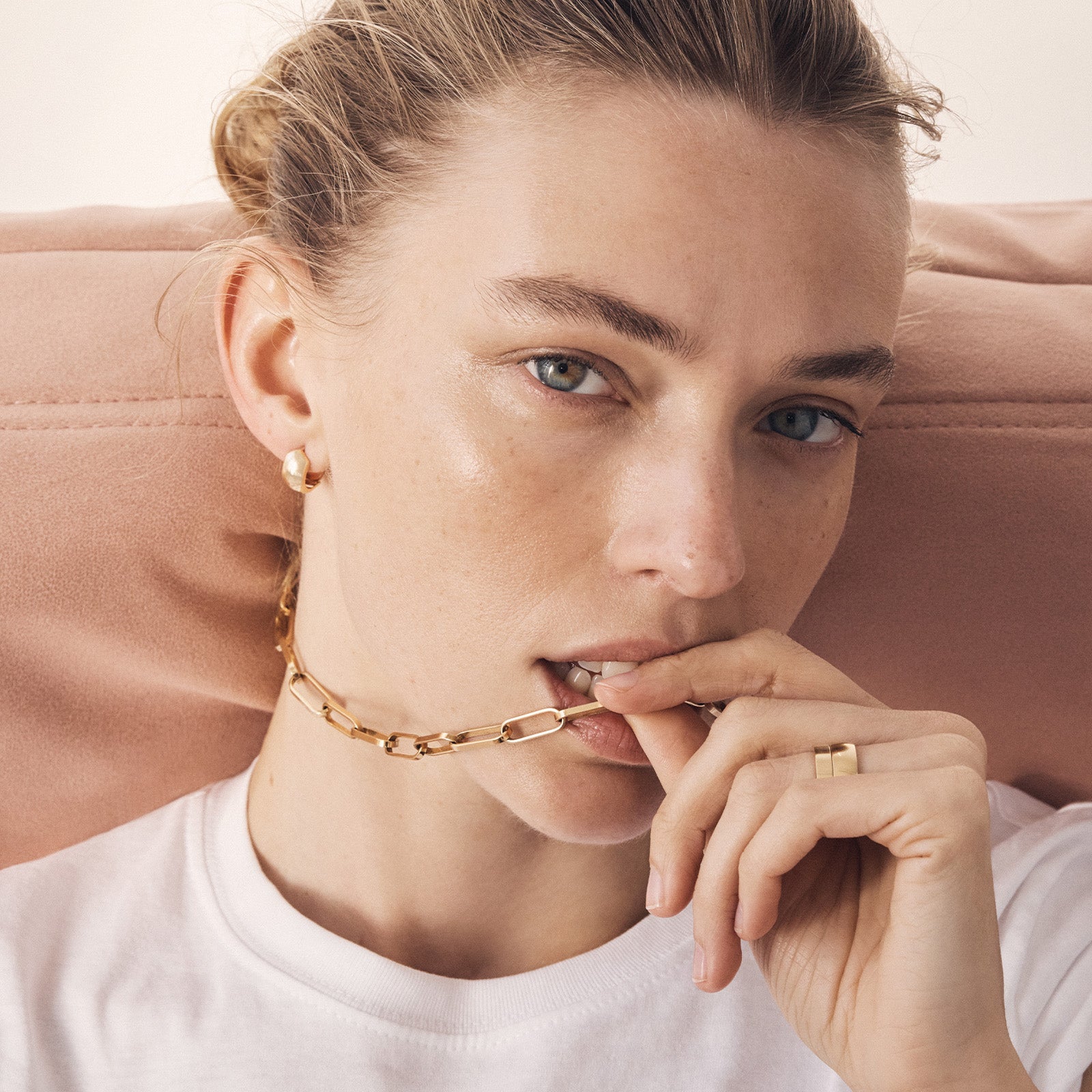 Woman wearing gold jewelry including earrings, a necklace, and ring, with a neutral background.