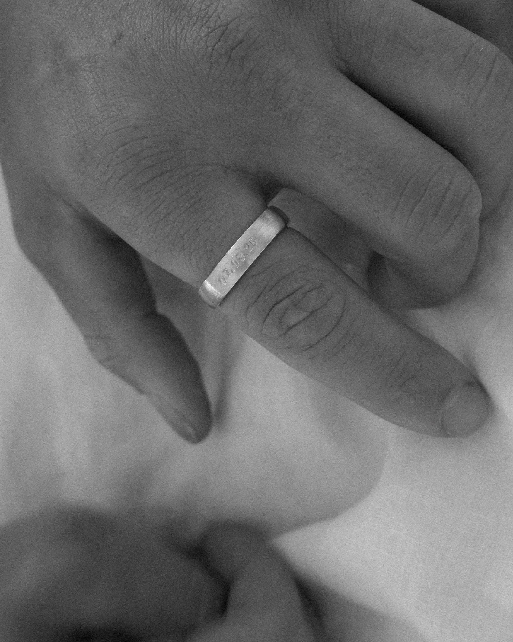 A close-up black and white photograph of a person's hand wearing a simple metal band ring on one finger, resting on a soft white fabric.