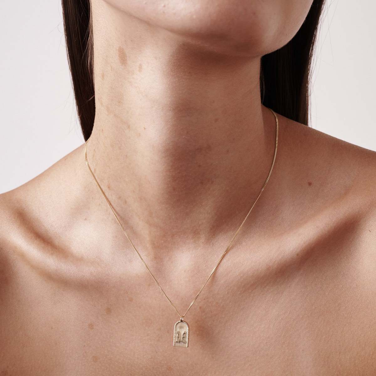Close-up of a woman wearing a delicate gold necklace with a rectangular pendant featuring an engraved arch design on her neck.