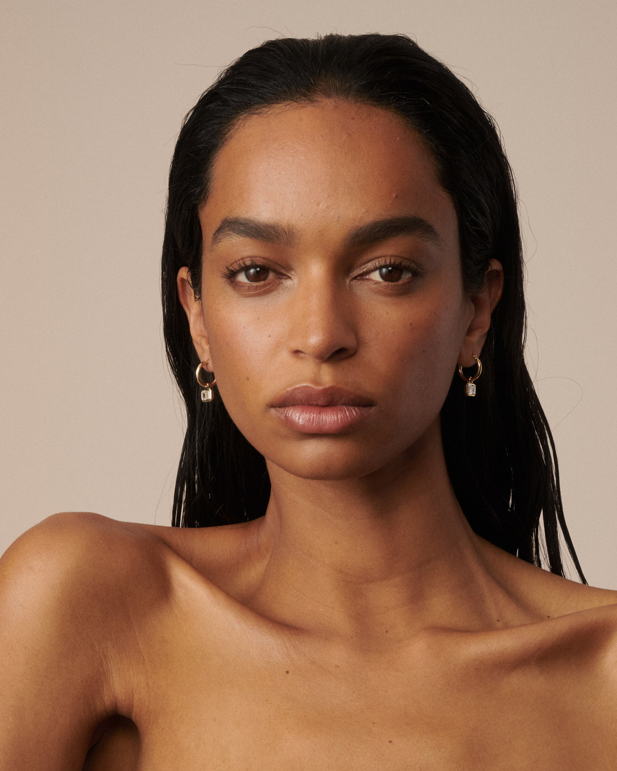A close-up portrait of a woman with dark hair wearing small gold hoop earrings with clear diamond drops, set against a solid beige background.