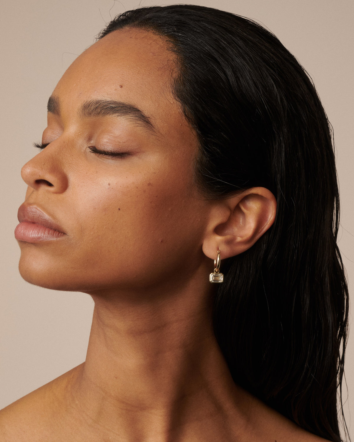 A close-up side profile of a woman with dark hair wearing a small gold huggie hoop earring with a clear rectangular gemstone, set against a solid beige background.