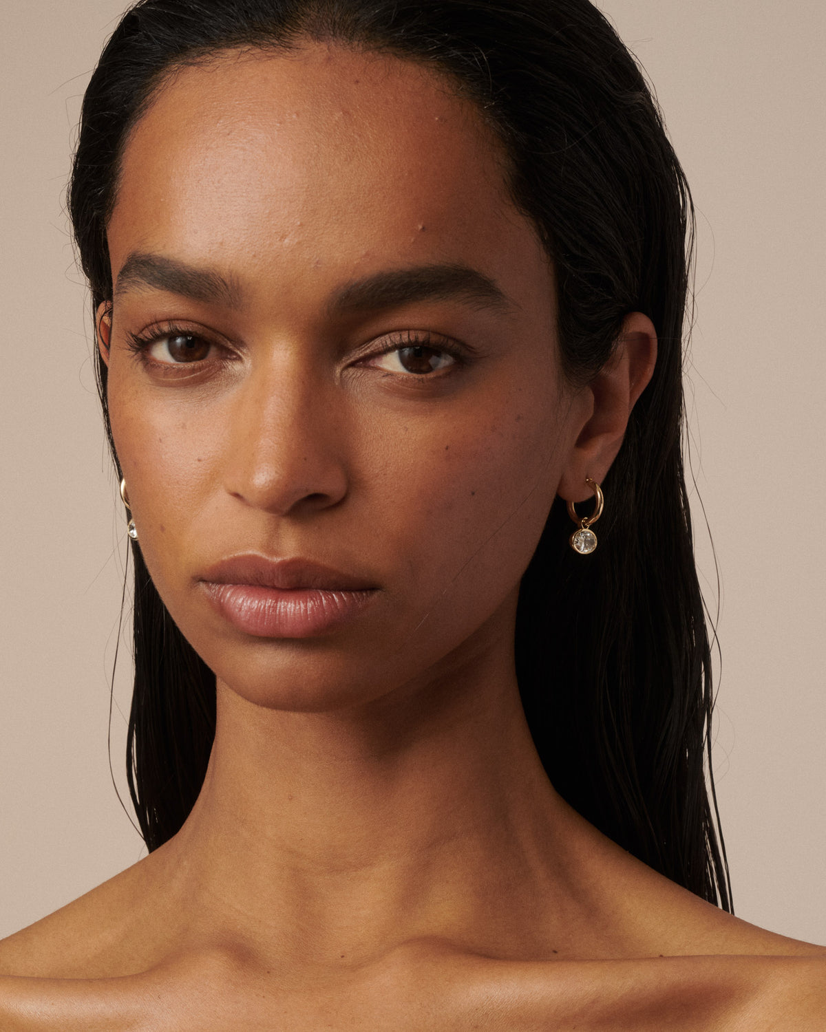 A close-up portrait of a woman with dark hair wearing small gold hoop earrings with clear diamond drops, set against a solid beige background.