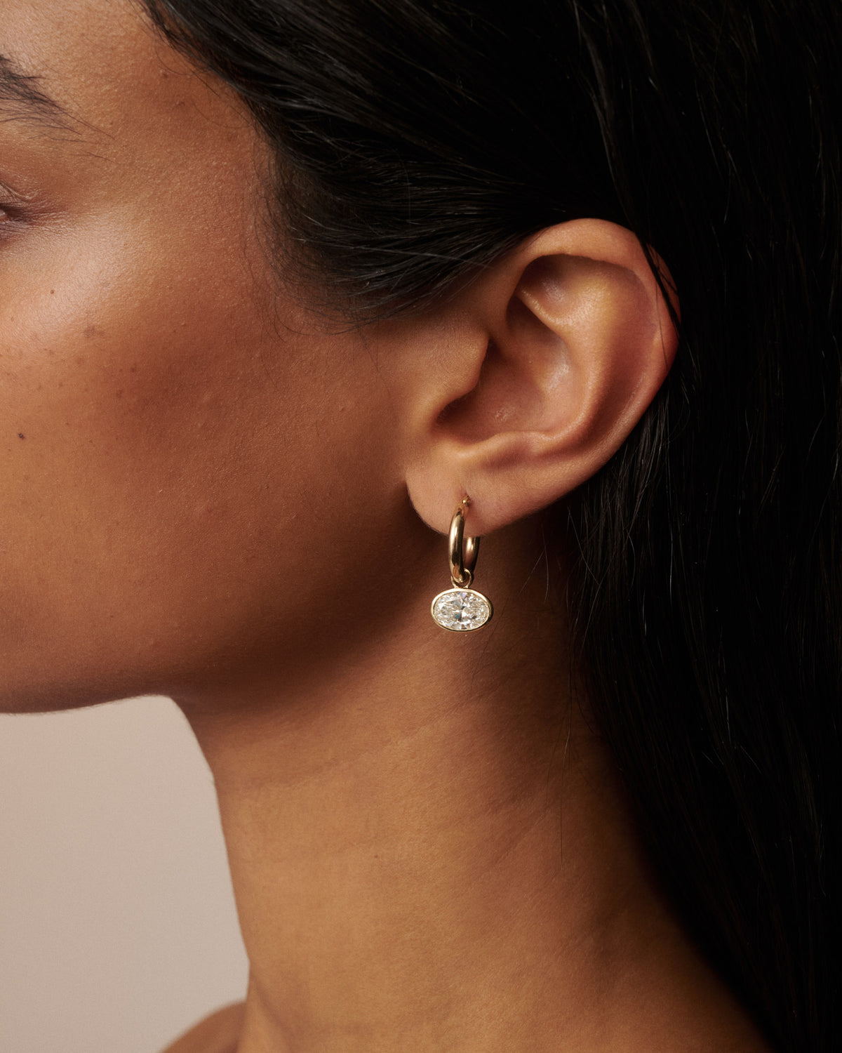 A close-up portrait of a woman with dark hair wearing small gold hoop earrings with clear diamond drops, set against a solid beige background.