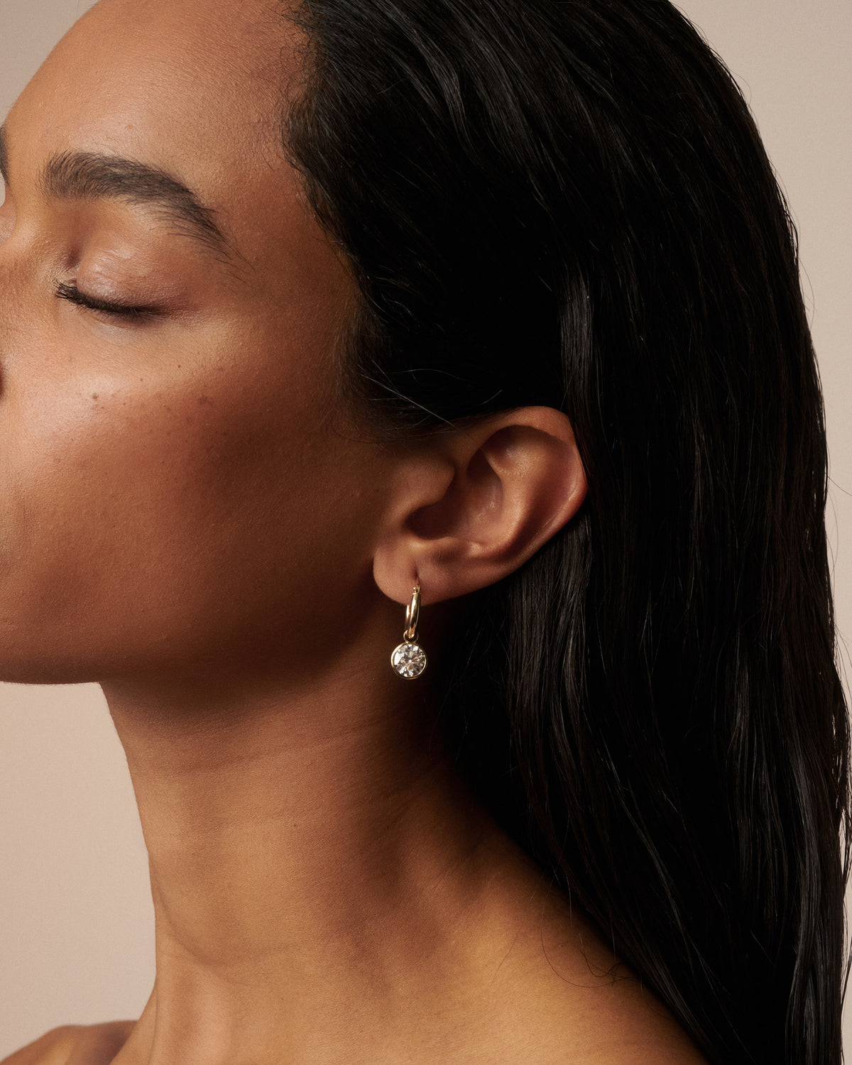 A close-up side profile of a woman with closed eyes wearing a delicate gold hoop earring with a small clear faceted drop stone, set against a warm beige background.