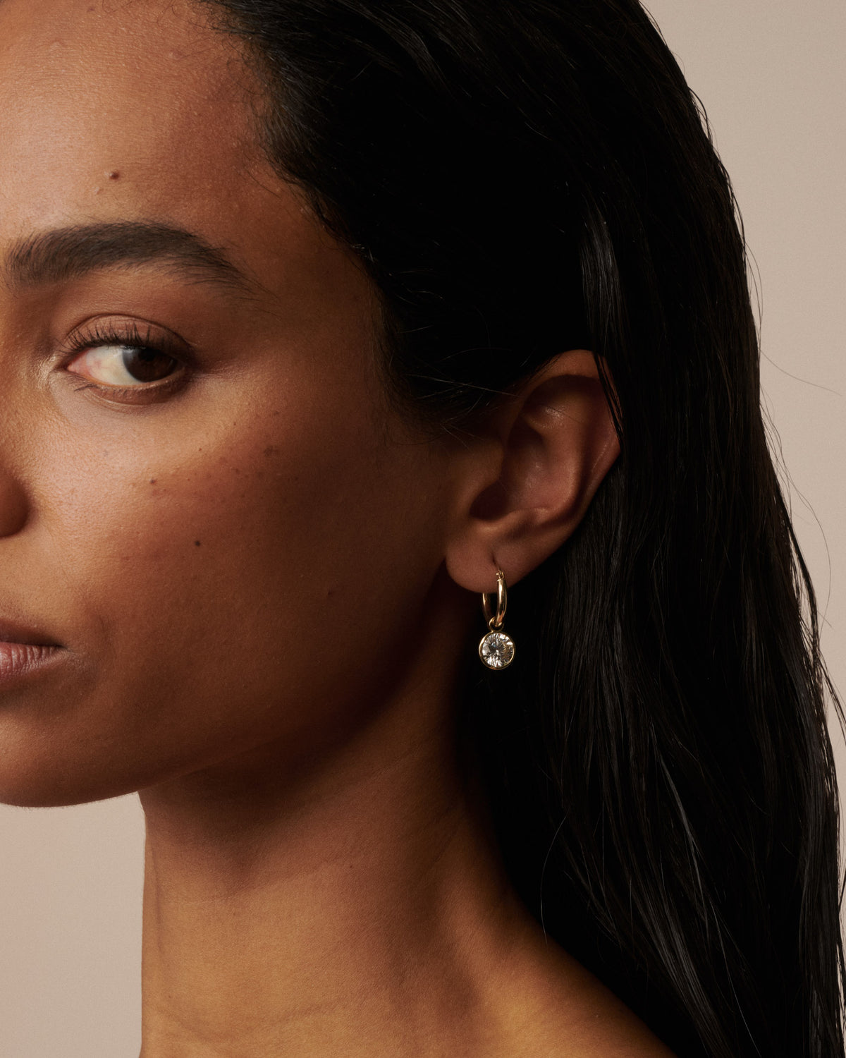 A close-up of a woman's ear wearing a delicate gold hoop earring with a small clear round drop pendant, set against a warm beige background.