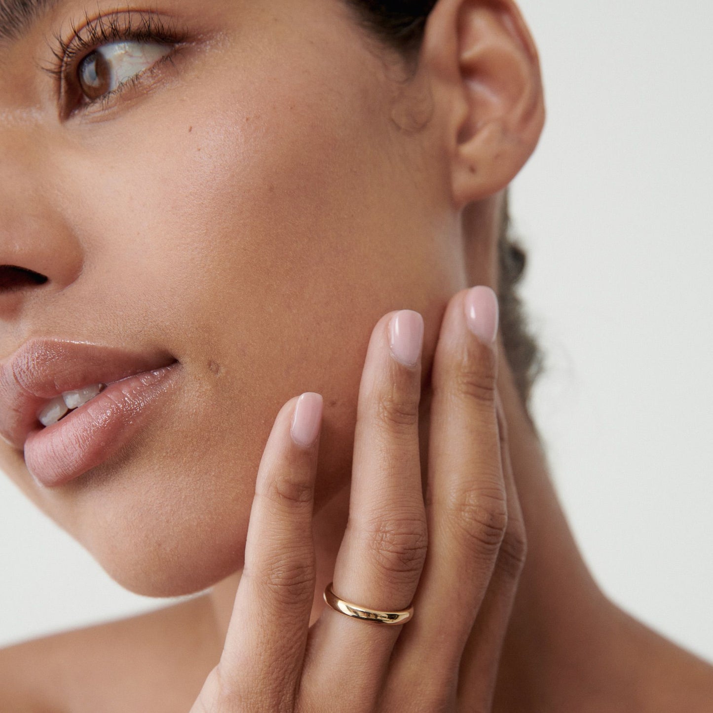 Close-up of a woman's face and hand, showcasing a minimalist gold ring on her finger, with a soft pink nail polish. Luxe jewellery detail.