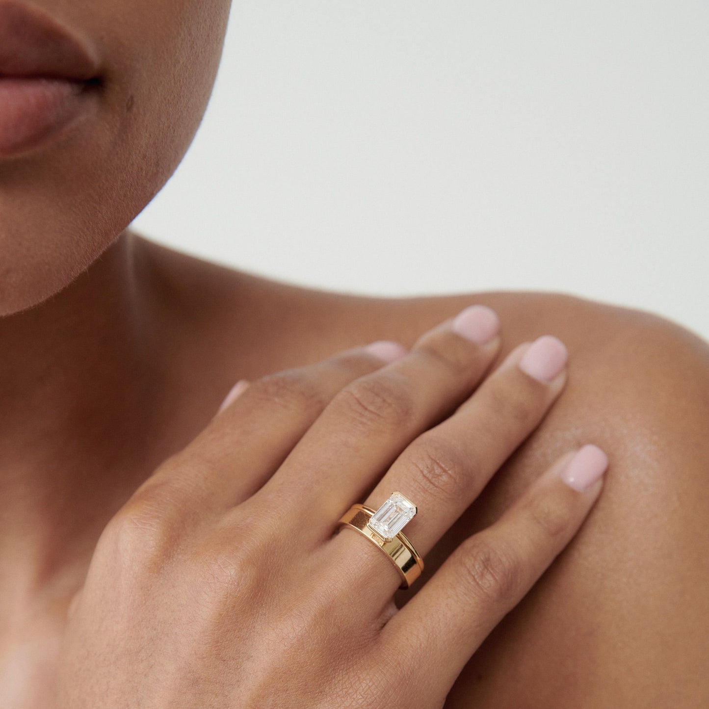 Close-up of a woman’s hand showcasing an emerald-cut diamond ring on a gold band, elegantly displayed against her shoulder.  [text overlay: Medium and 1ct Rya]