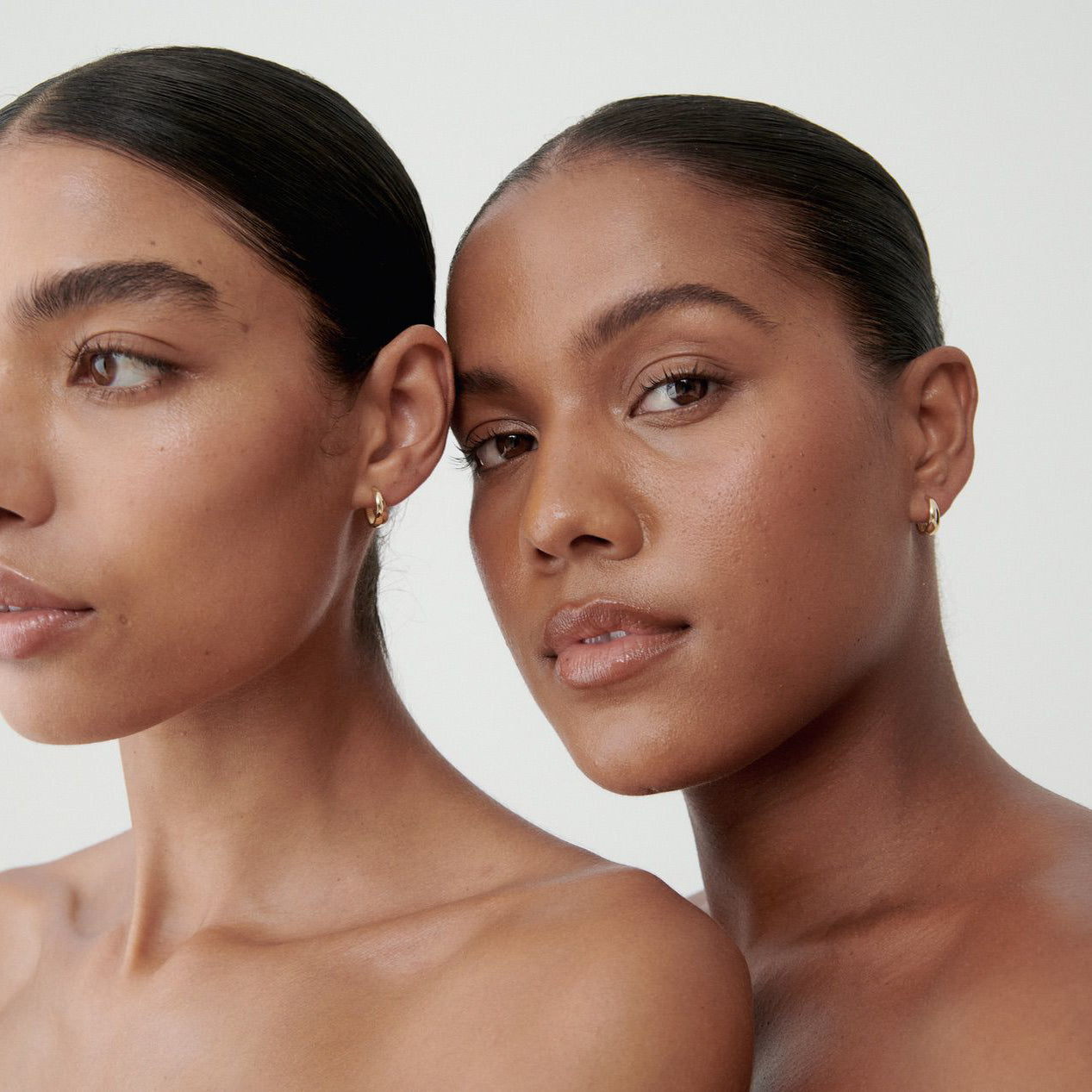 Two women with sleek hair showcase minimalist gold hoop earrings against a neutral background, emphasising luxury jewellery elegance.