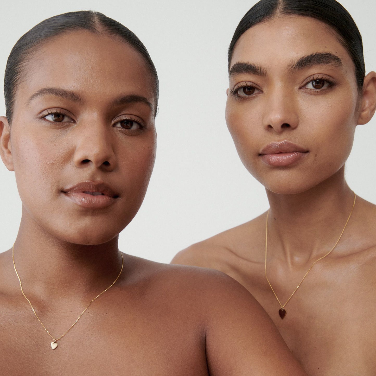 Two women wearing elegant gold necklaces with heart pendants; bare shoulders and neutral backgrounds emphasise luxury jewellery design. [text overlay: Small pendant (left) and large pendant (right)]

