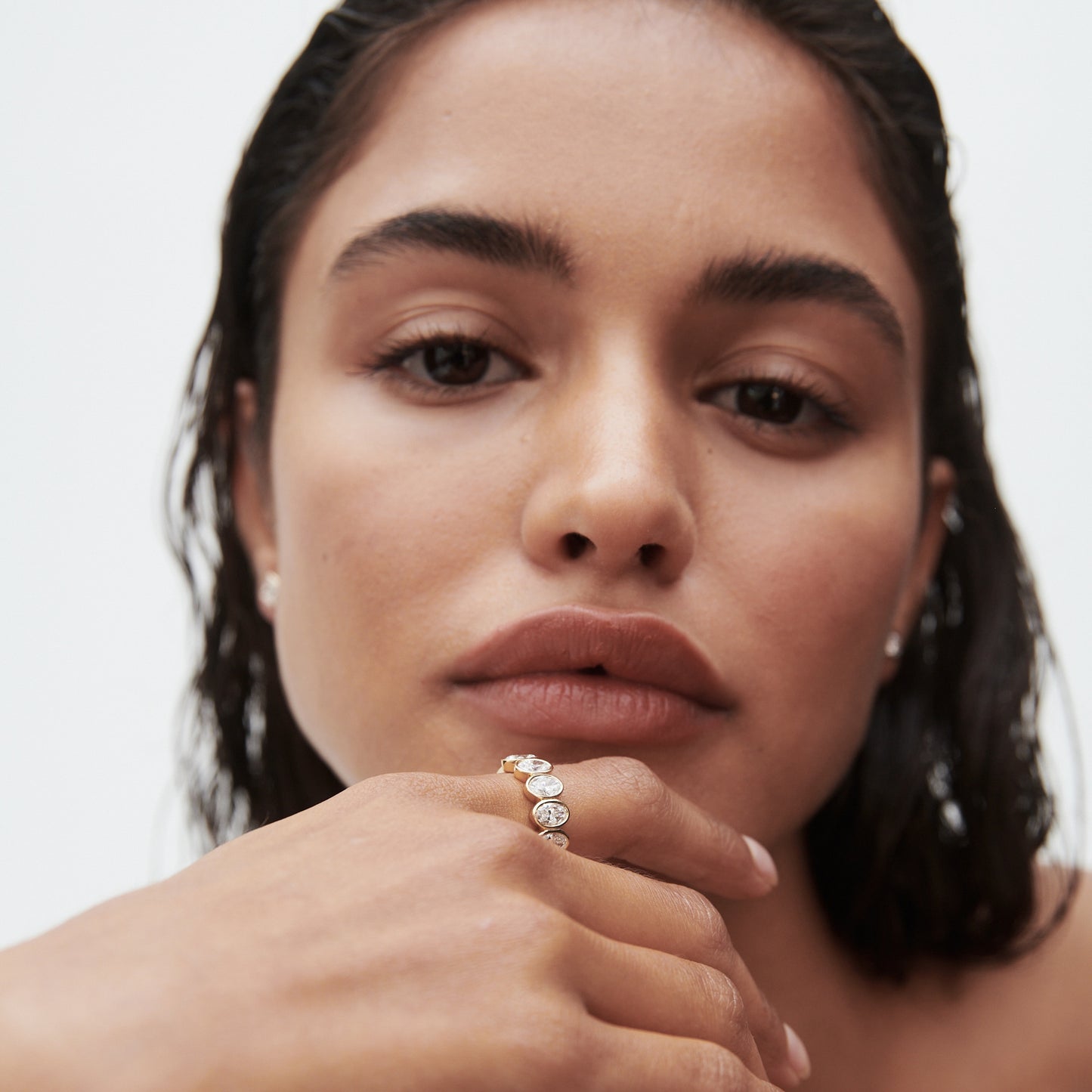 Woman with wet hair showcasing a luxurious multi-stone ring, earrings, and natural makeup against a plain background.
