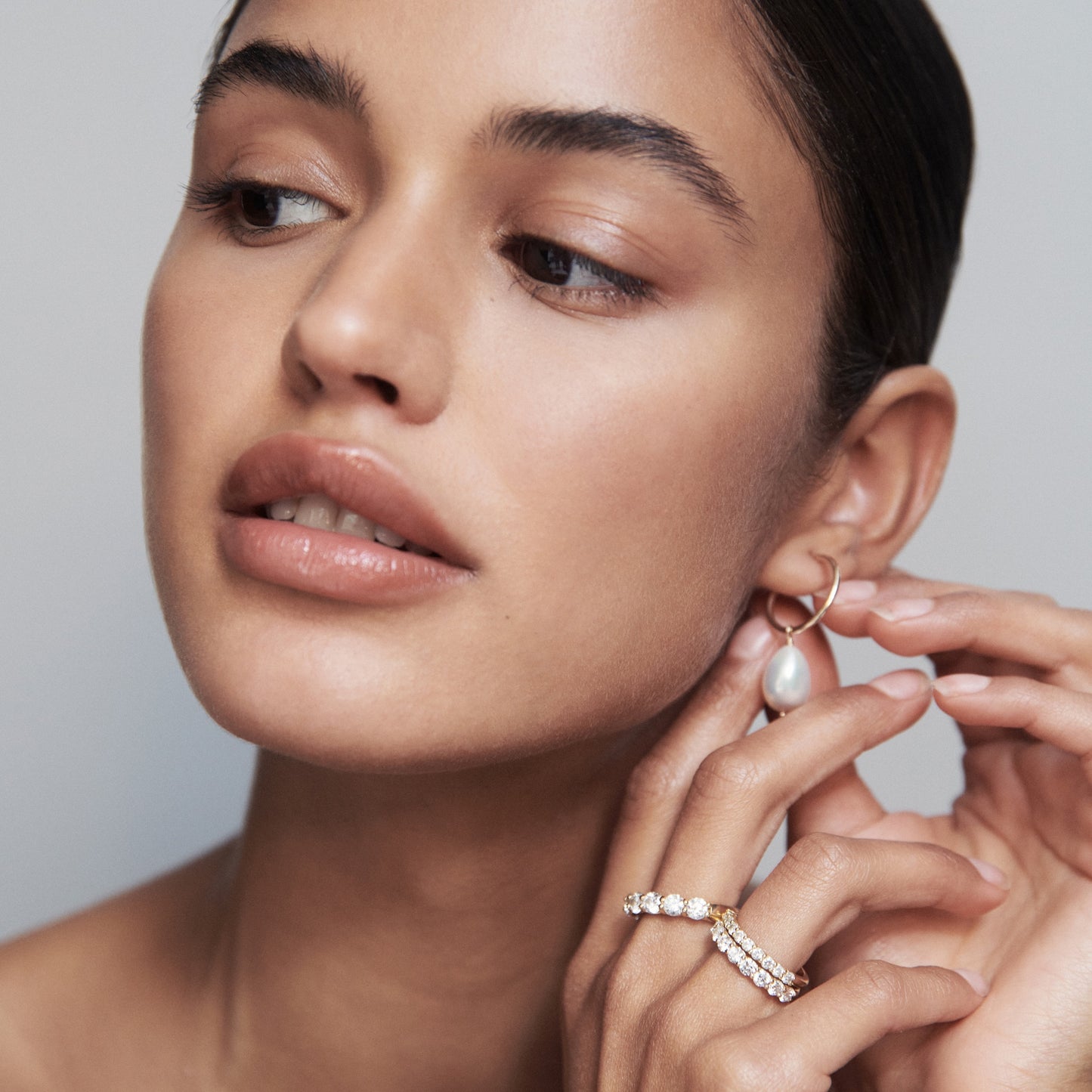 Woman wearing gold hoop earrings with a dangling pearl and a gold diamond ring; showcasing luxury jewellery. [text overlay: Pictured with Arraya Diamond Bands]