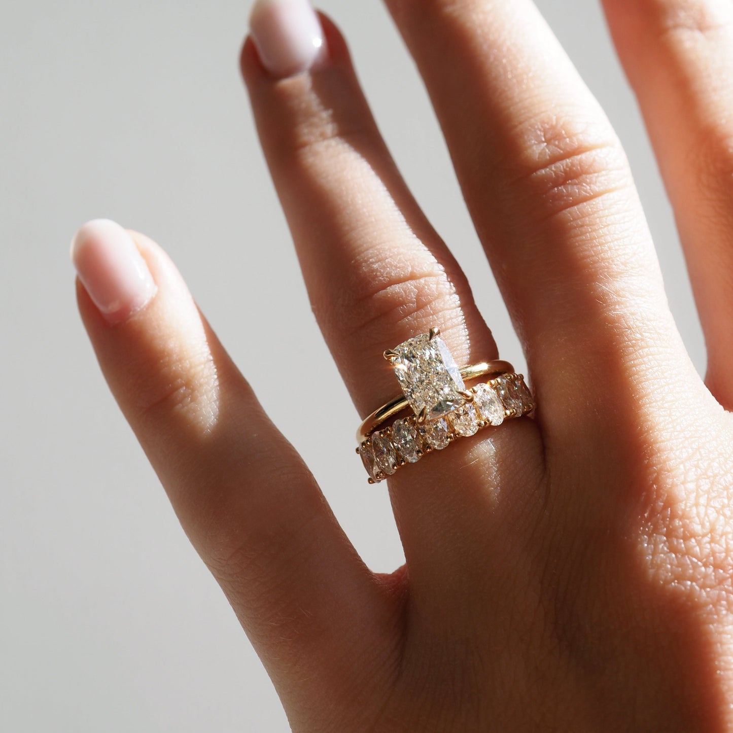 Close-up of a hand wearing two gold rings with radiant-cut diamonds, showcasing luxury jewellery against a soft, natural light background.[text overlay: 2ct Diamond and Camille Diamond Ring]
