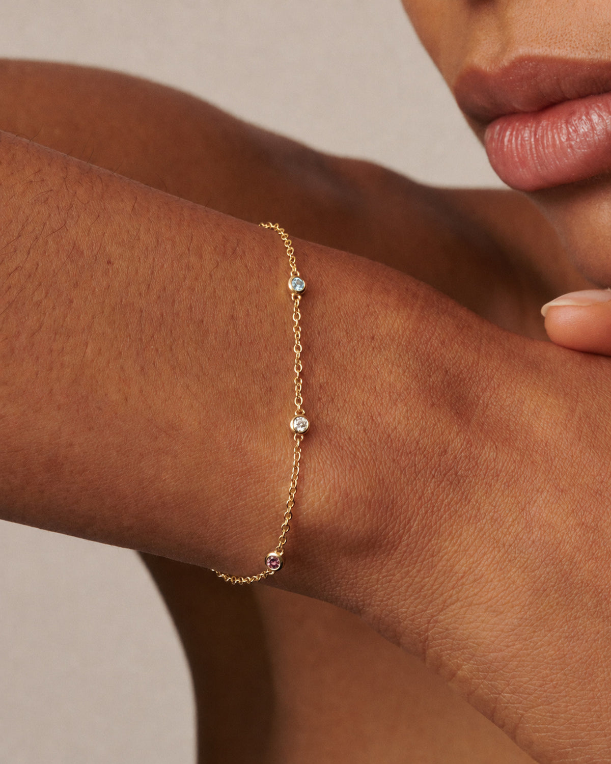 A close-up of a model's wrist wearing a delicate gold chain adorned with small clear gemstones.