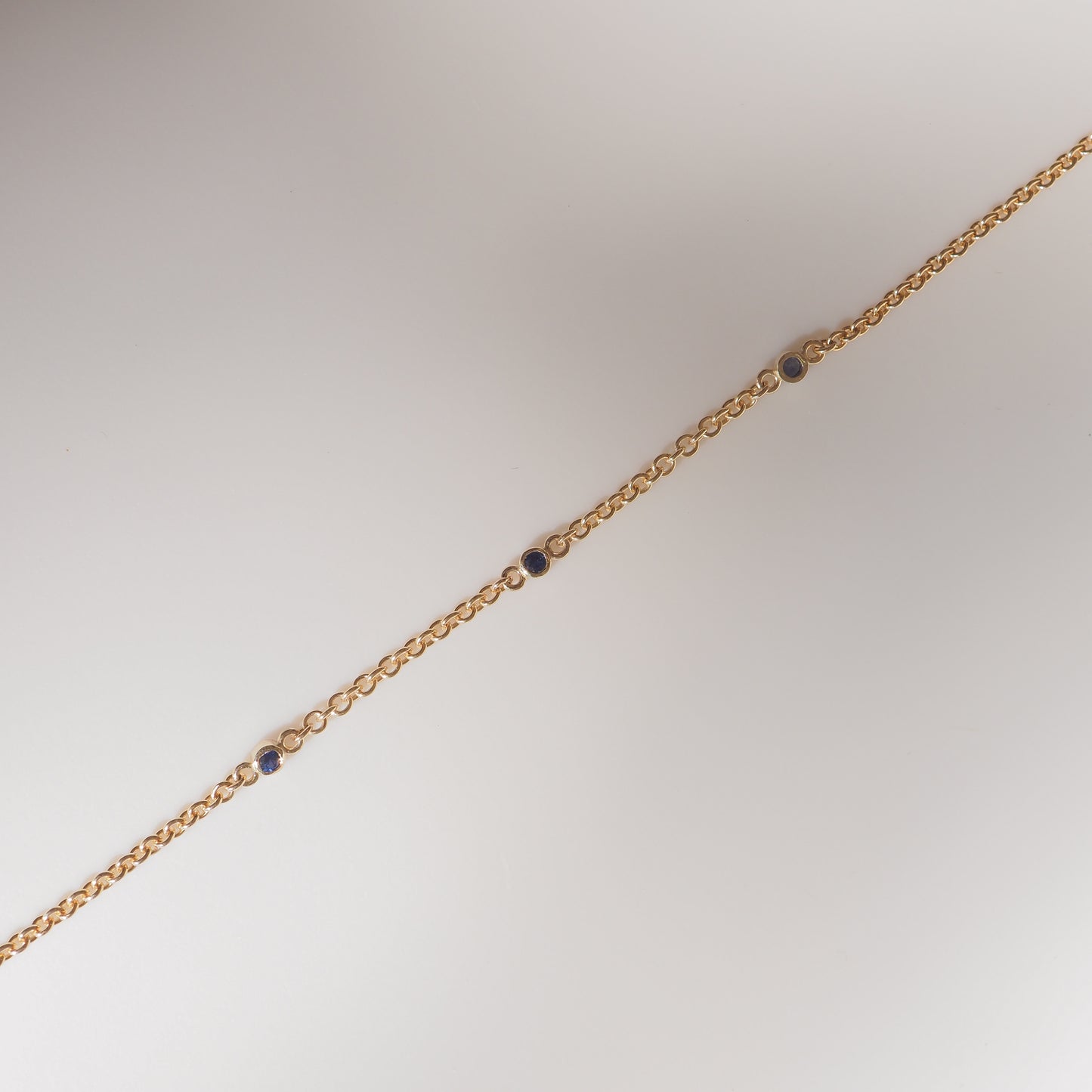 Gold chain bracelet with round links and scattered blue gemstones on a light background. Luxurious jewellery design.