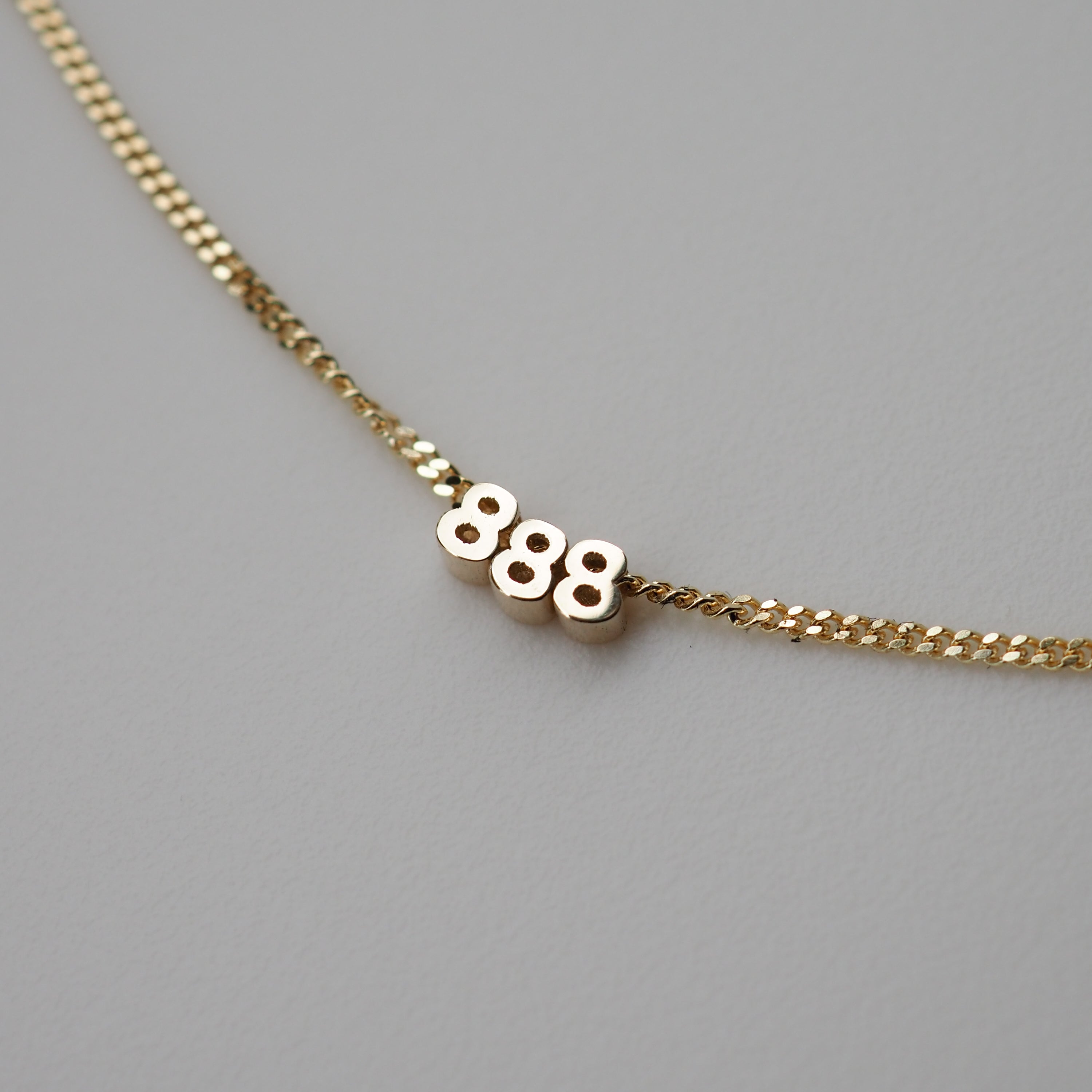 Elegant gold necklace featuring the number "888" in bold on a fine chain, displayed against a plain grey background.