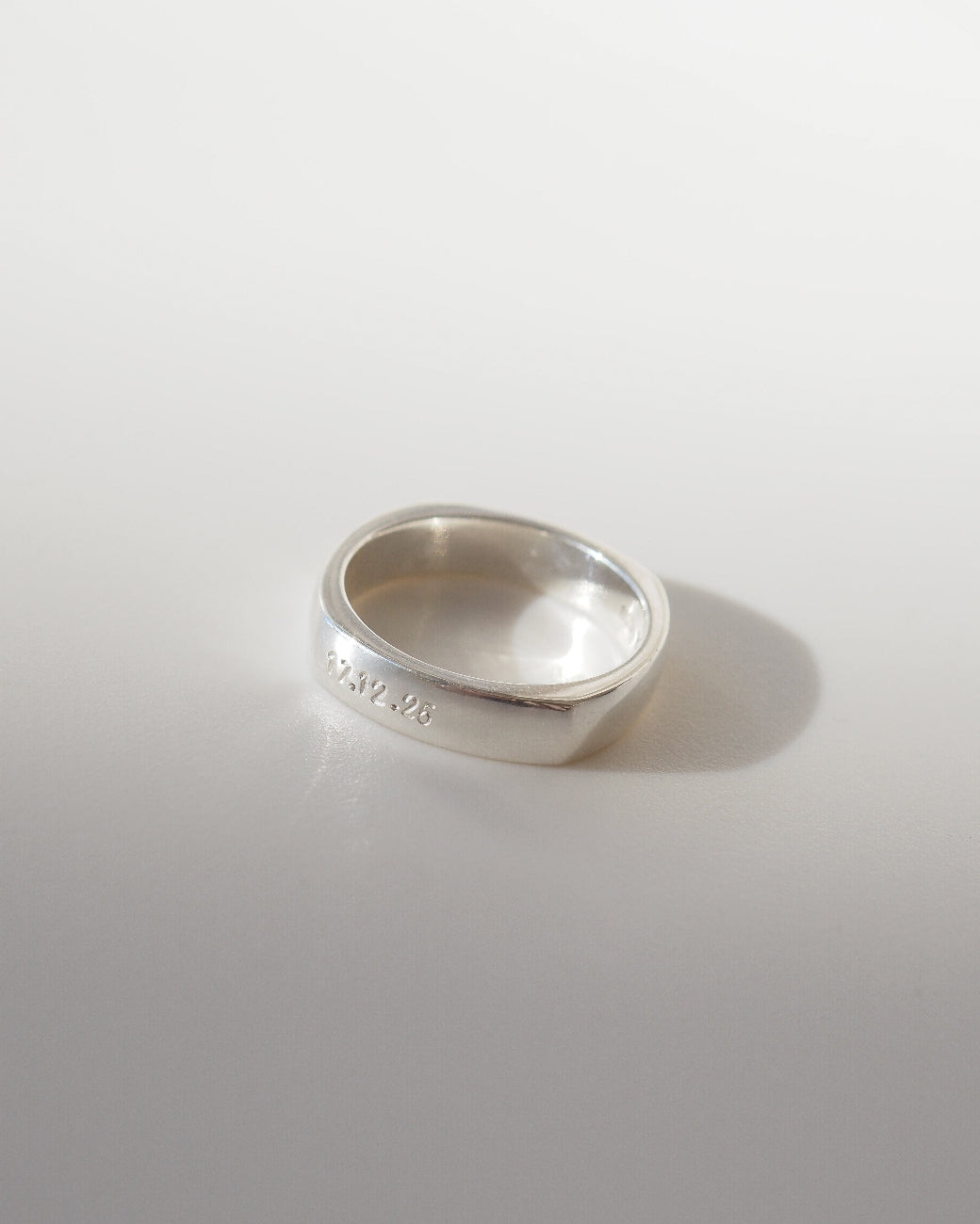 A polished silver band ring with engraved markings, resting on a light gray surface with a soft shadow.