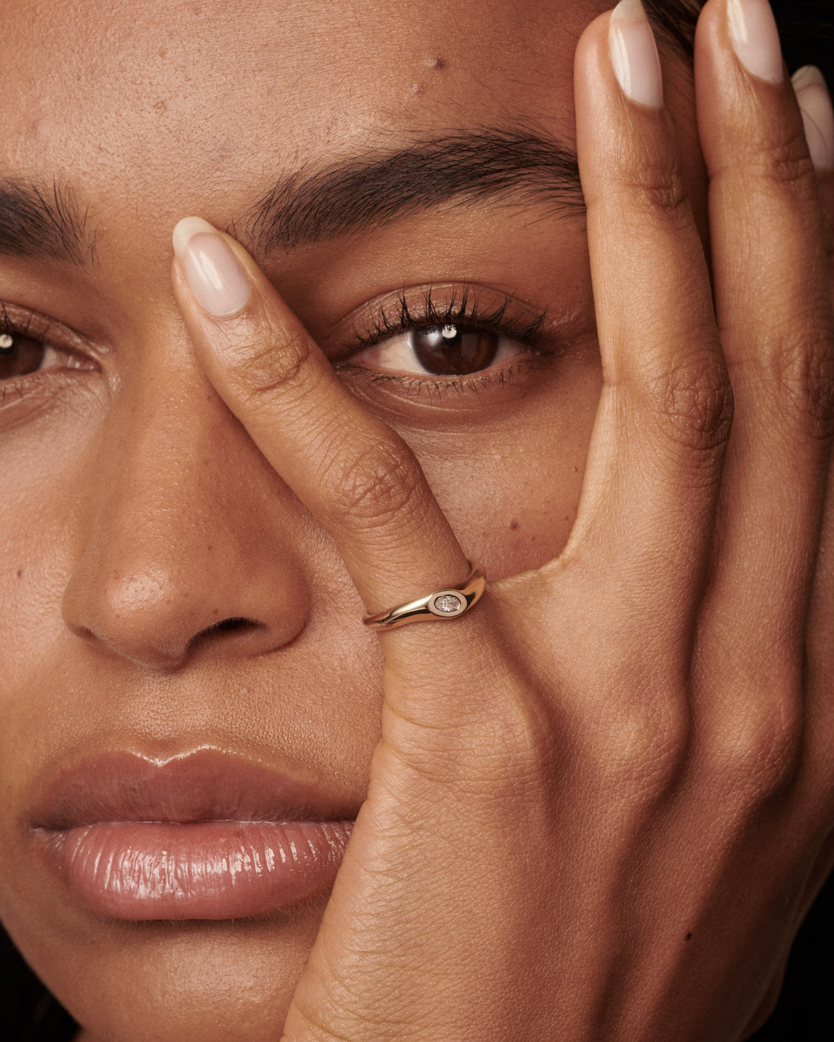 A close-up lifestyle shot of a model's face and hand, wearing a delicate gold ring with a small oval clear diamond on the ring finger. [text overlay: Francielle wears the 0.15ct]