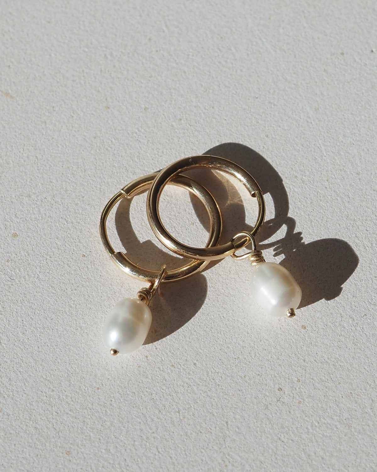 Gold hoop earrings with single dangling white pearls, casting soft shadows on a light surface. Luxurious jewellery design.