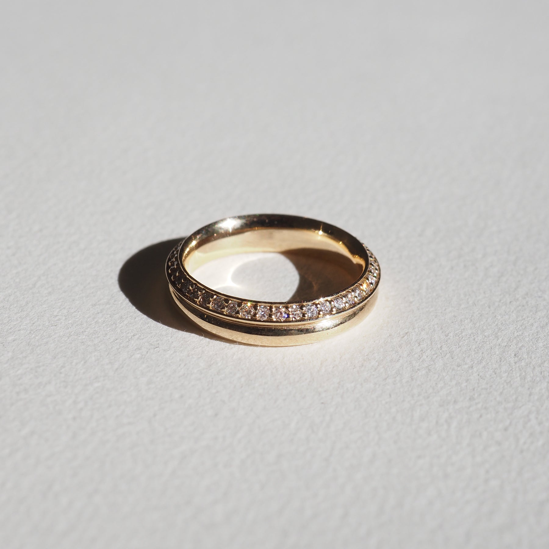 Gold ring featuring a row of embedded diamonds, casting a soft shadow on a light surface. Elegant and luxurious jewellery piece.