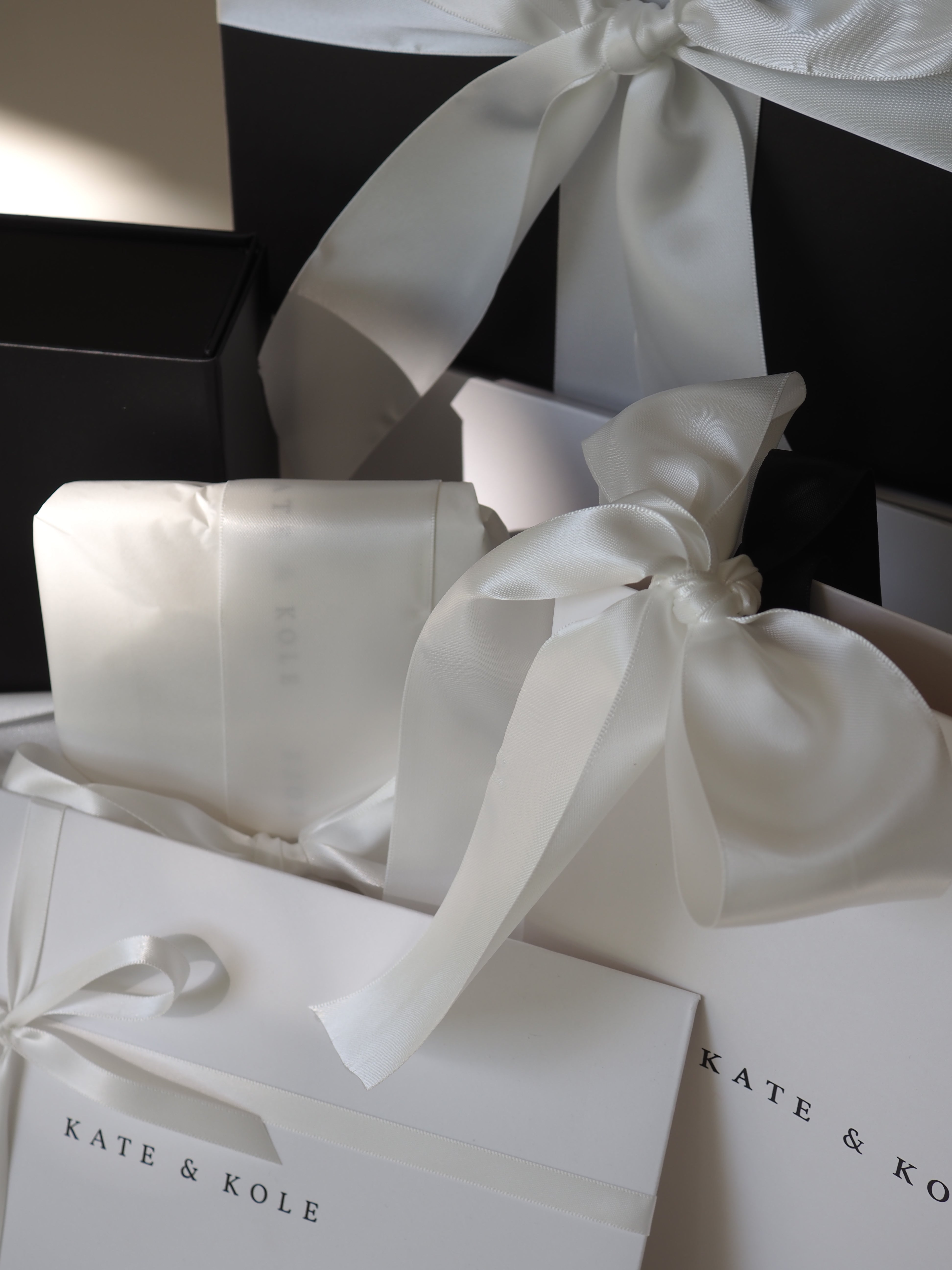 Luxurious jewellery packaging with elegant white satin bows and black boxes, featuring the brand name "Kate & Kole" for a sophisticated impression.