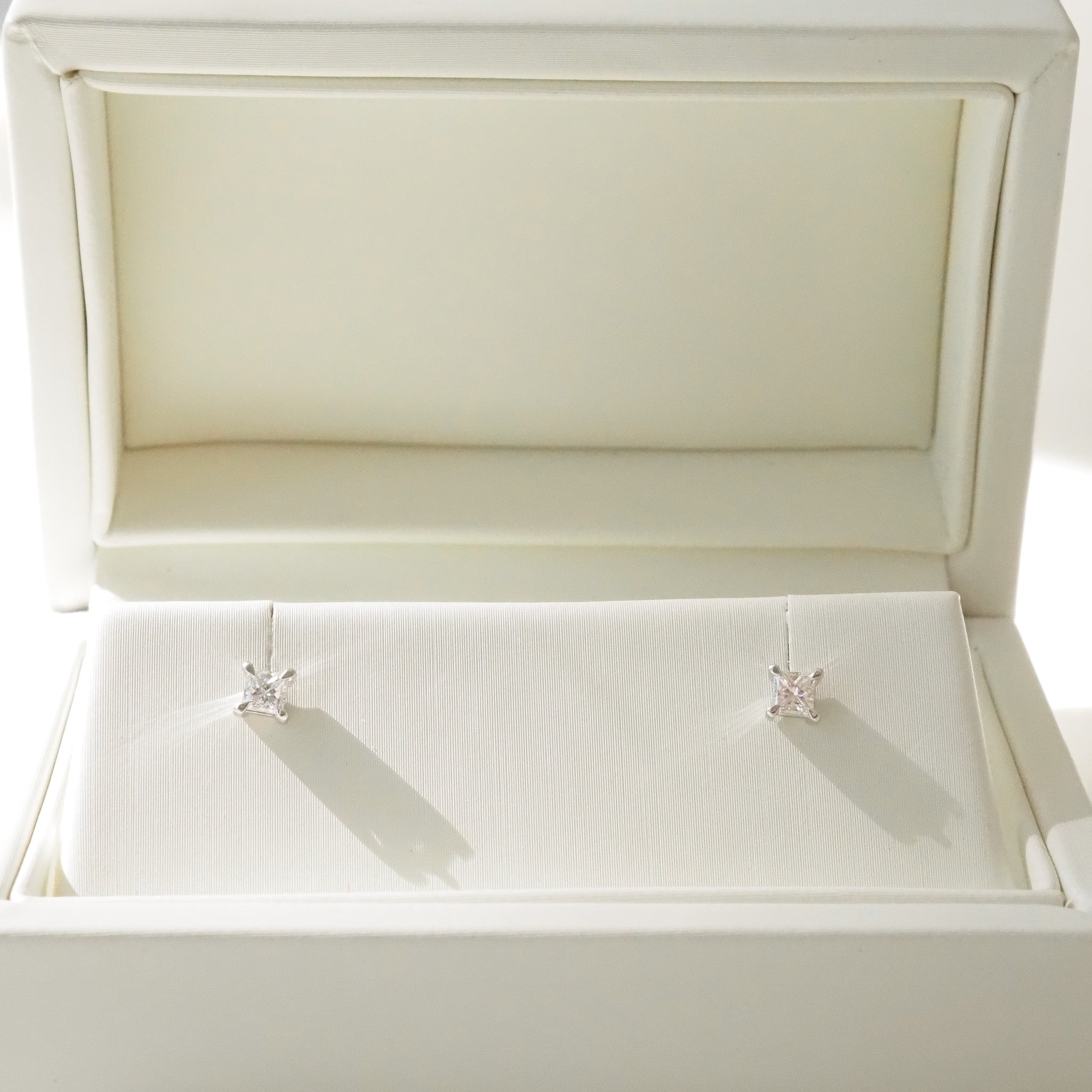 Pair of princess diamond stud earrings in an open white jewelry box.