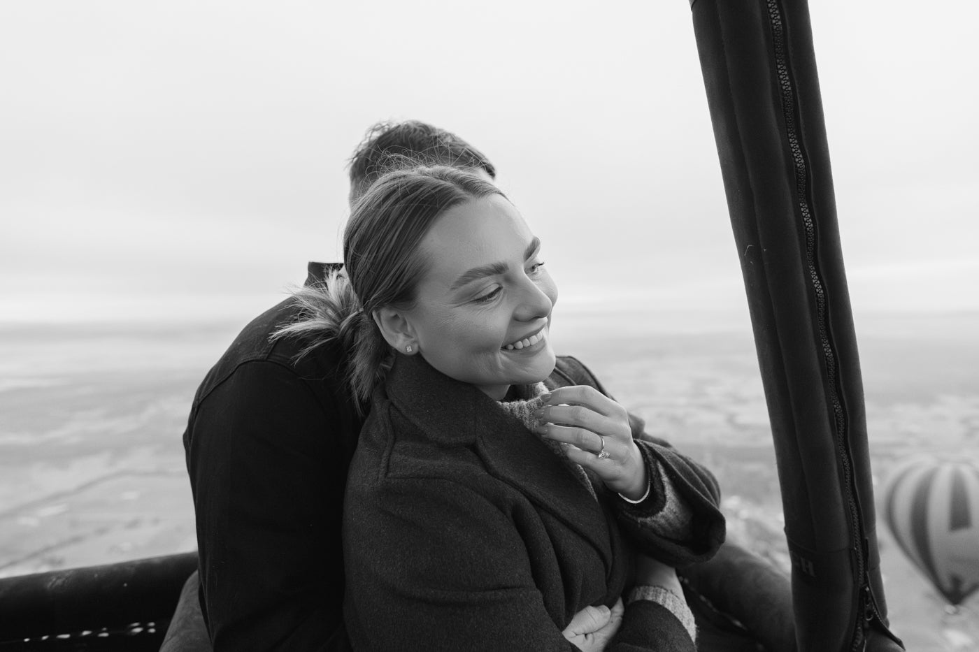 Couple enjoying a hot air balloon ride, woman smiling with a hand on her chest, showcasing an elegant engagement ring. Scenic landscape backdrop.