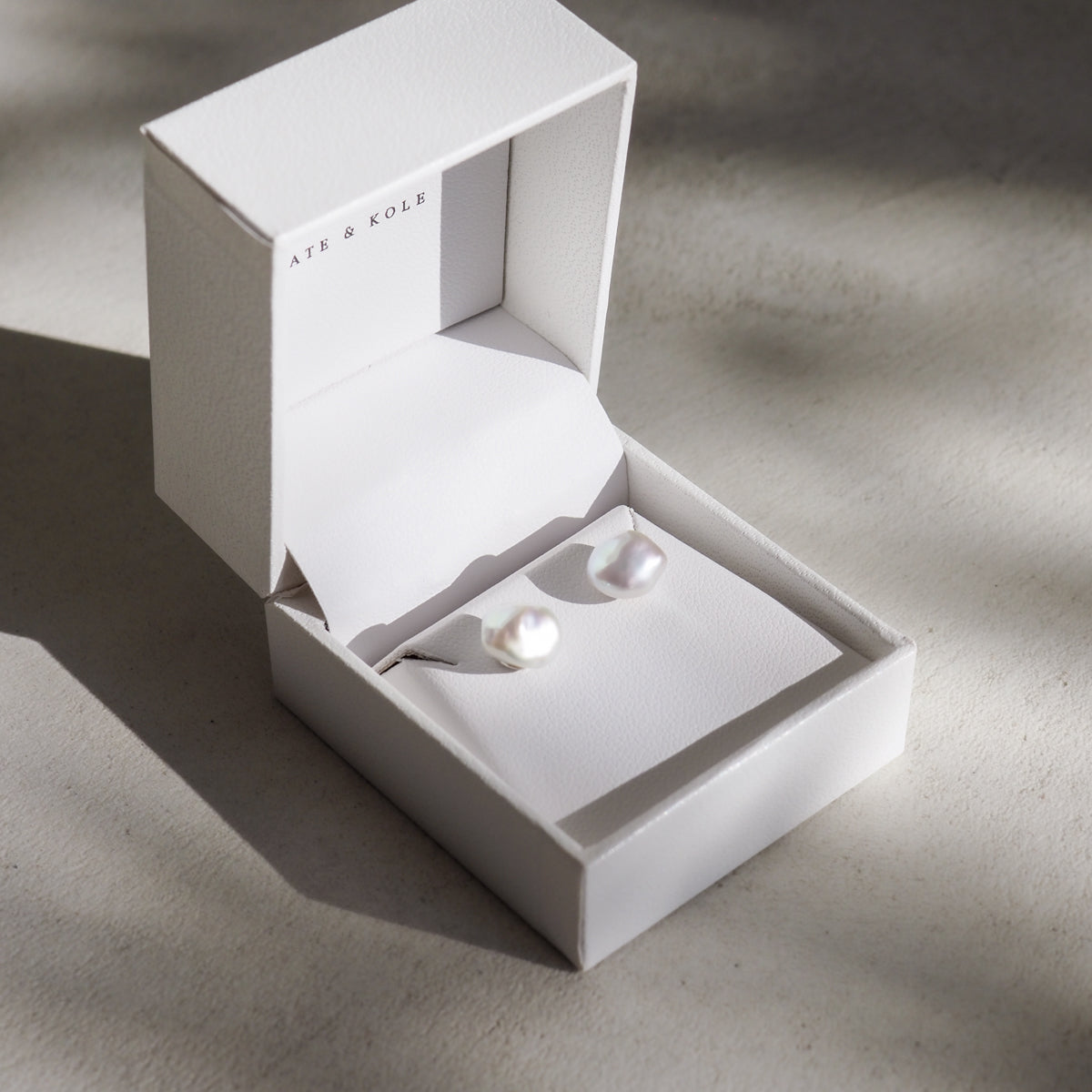 White pearl earrings in a white open jewellery box on a light grey background. Shadows add elegance, highlighting the luxury jewellery piece.
