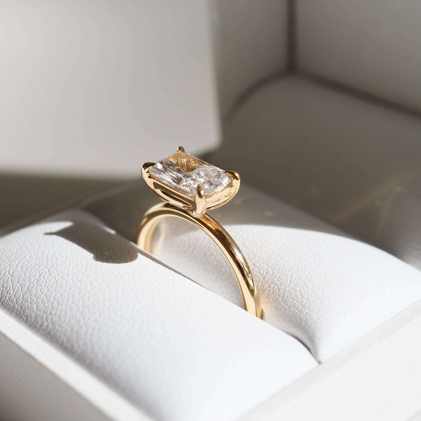 Elegant gold ring featuring a radiant-cut diamond, displayed in a luxurious white jewellery box under soft lighting.