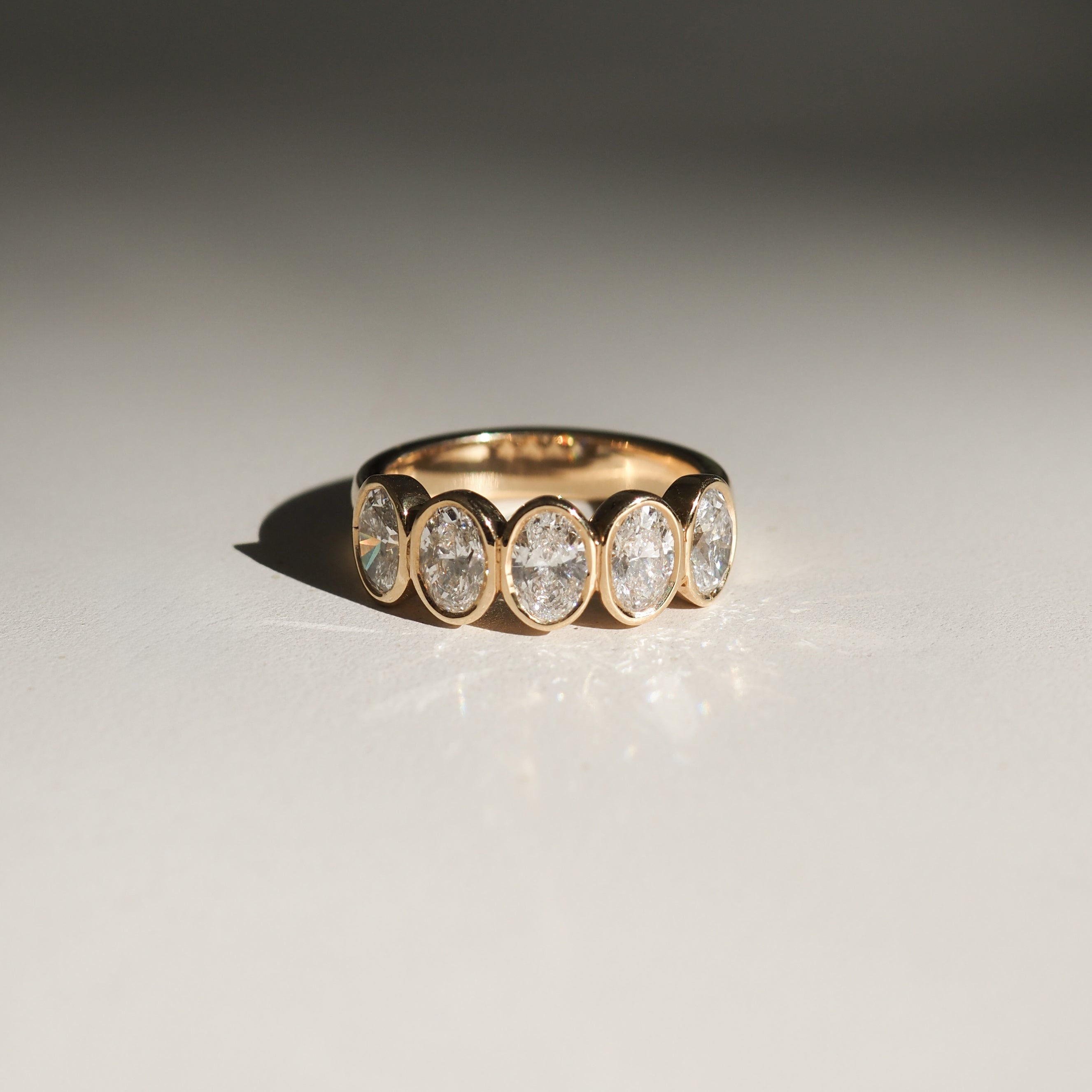 Gold ring with five oval diamond settings, displayed on a reflective surface. Elegant and luxurious jewellery design.