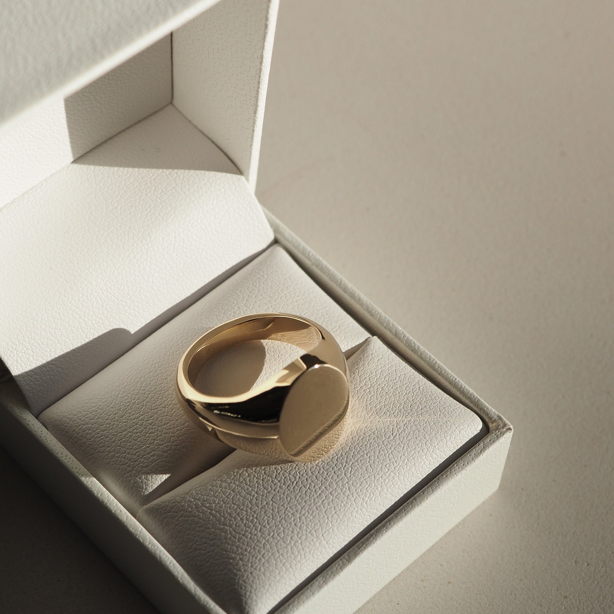 Gold signet ring in an open white jewellery box, casting a subtle shadow on a light surface, showcasing luxury craftsmanship.