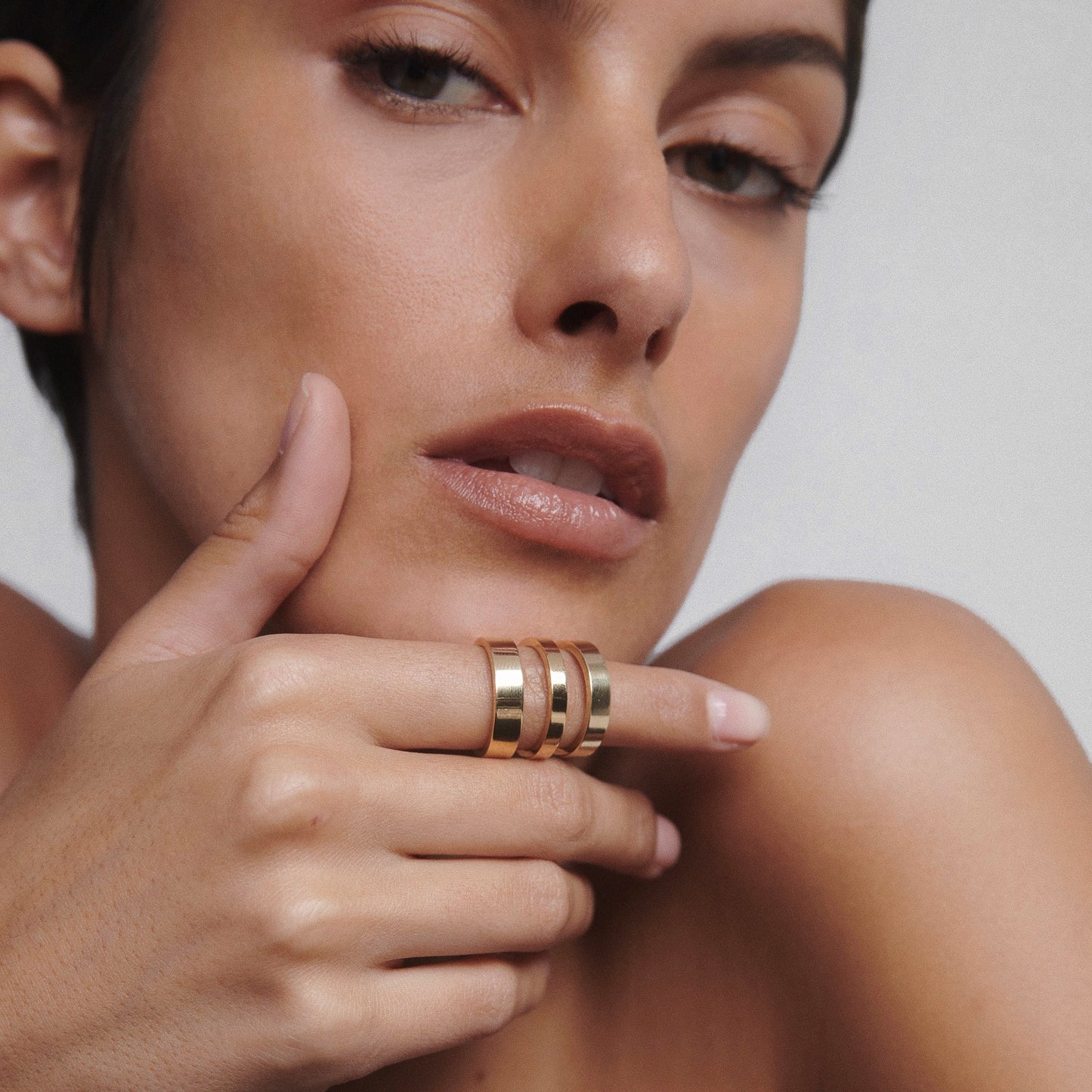 Woman's hand with luxurious, stacked gold rings held near her lips, showcasing elegant jewellery against a neutral background.  [text overlay: Bold (left), Fine (middle) and Medium (right)] 