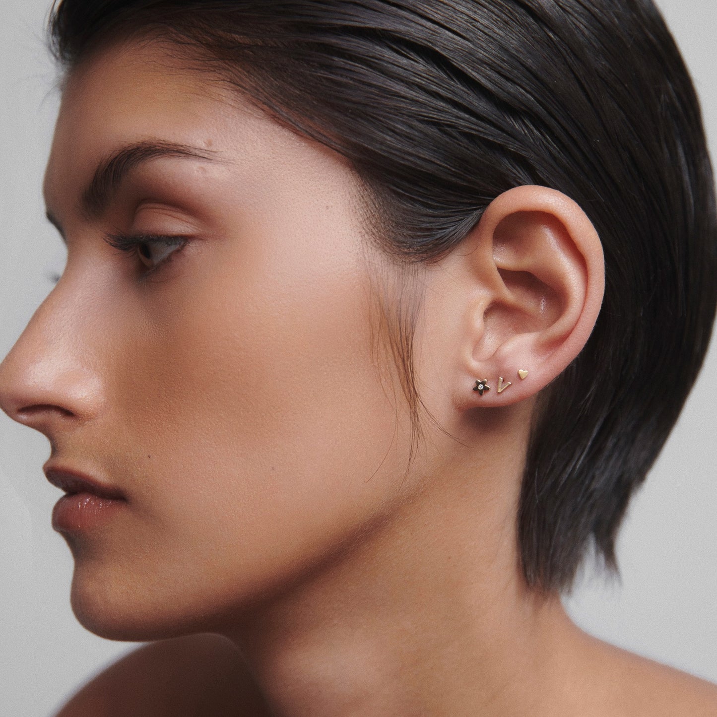 Profile of a woman with short hair wearing multiple gold and diamond earrings. Luxurious and elegant jewellery style. [text overlay: Flower Stud with diamond (left), Letter Stud (middle) and Micro Heart Stud (right) ]