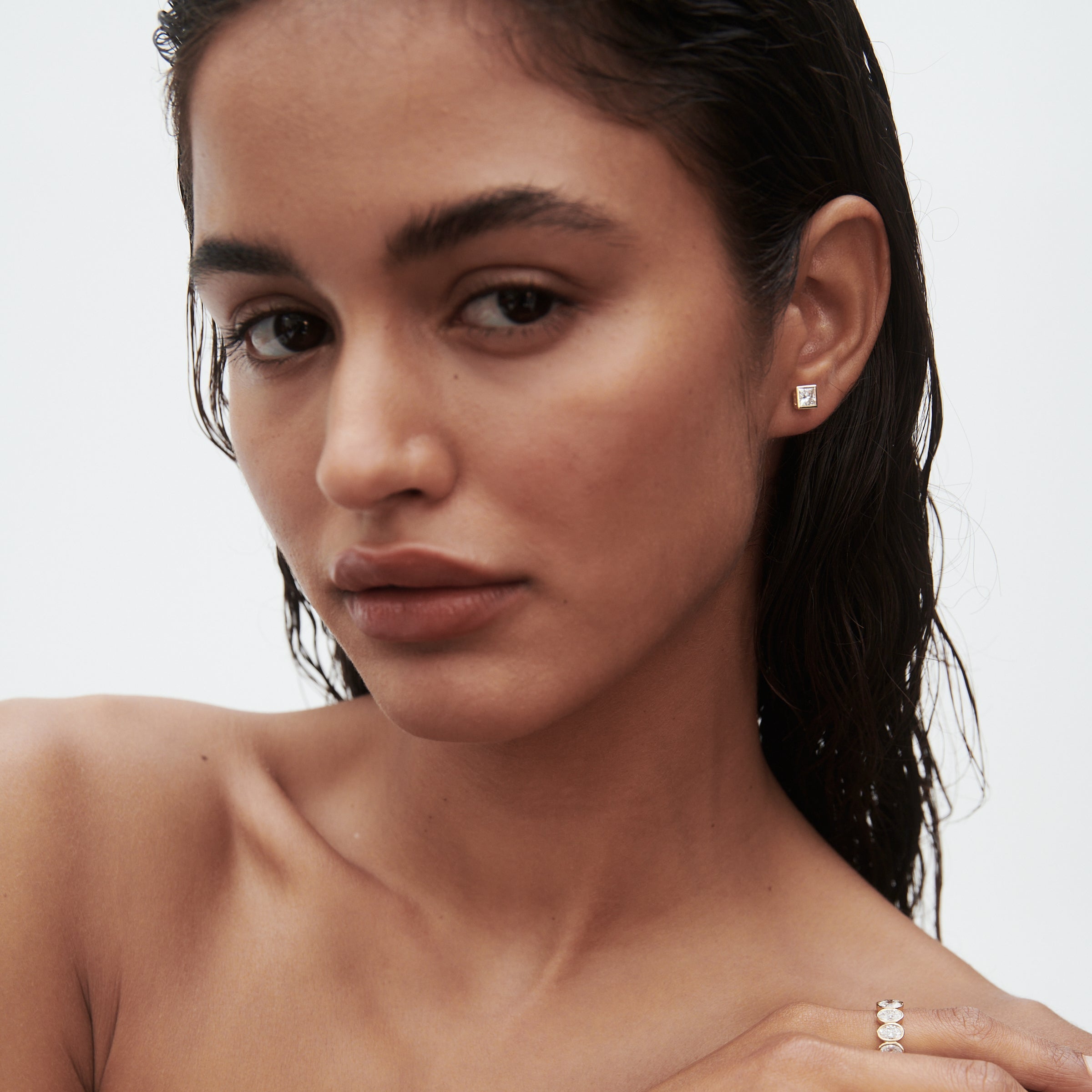 Woman with wet hair wearing square stud earrings and holding her chin, showcasing a ring with three gems. Sophisticated minimal jewellery style.