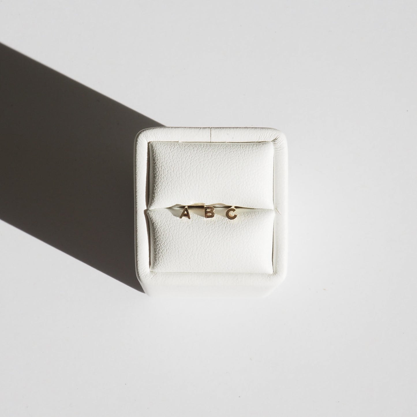 Gold initial bracelet with letters A, B, C on white cushion, casting shadow on white surface. Elegant and minimalist jewellery design.