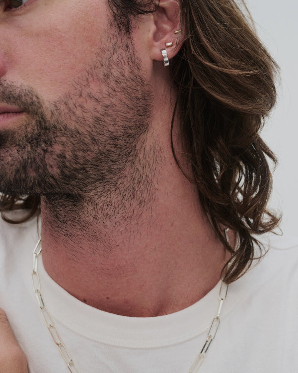 Close-up of a man's ear and neck showing silver hoop and stud earrings, along with a silver paperclip chain necklace, against a plain light background.