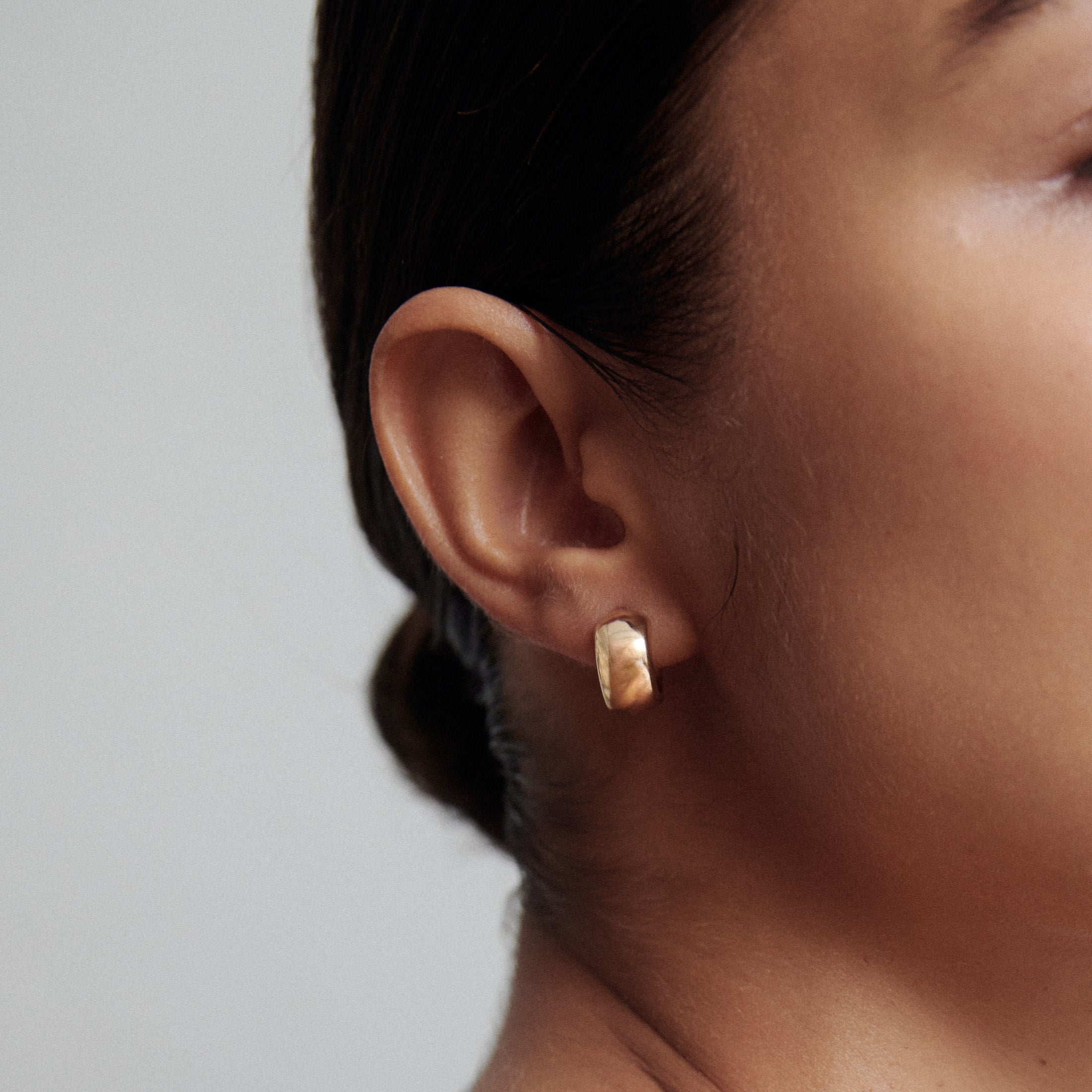 Close-up of a gold hoop earring on a woman's ear, showcasing luxury jewellery. The background is a neutral grey, highlighting the earring.