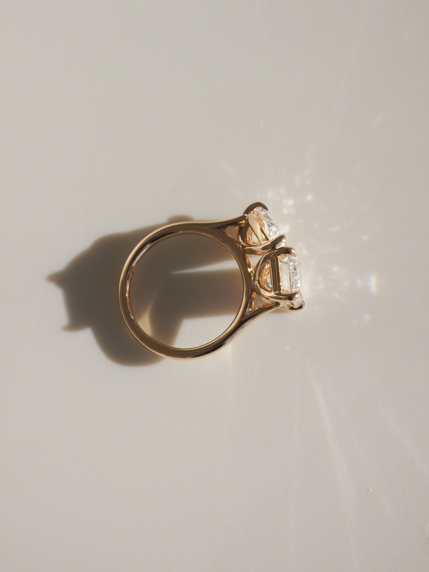 Gold ring with two sparkling emerald-cut diamonds casting a shadow on a beige surface, epitomising luxury jewellery elegance.