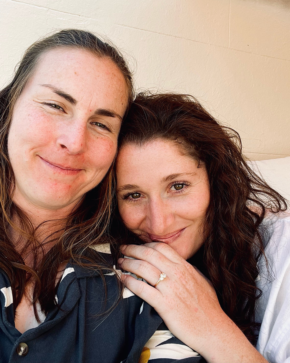 Two women smiling closely, one showing a sparkling engagement ring. Casual attire, warm setting, intimate moment.