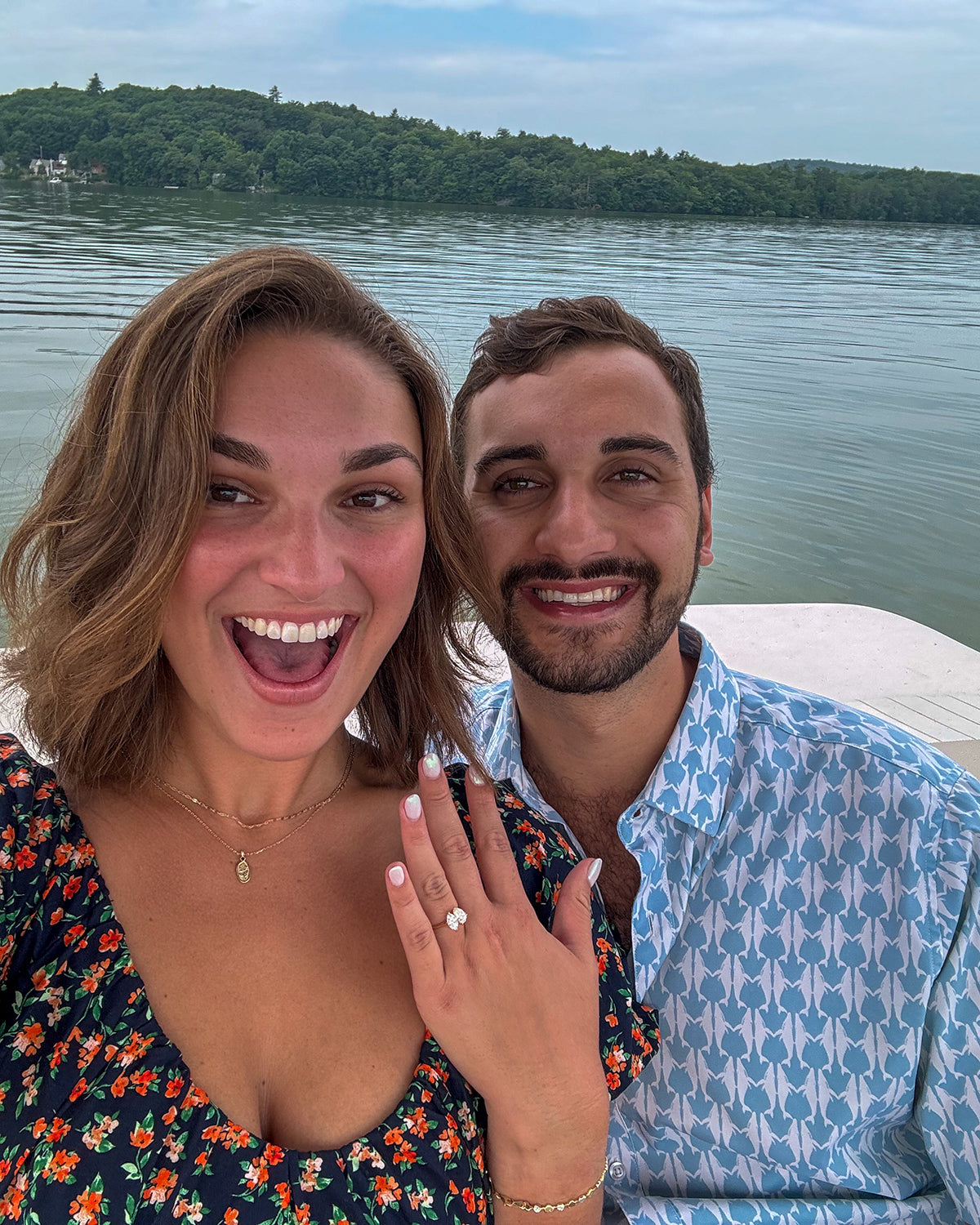 Couple smiling by a serene lake; woman prominently displaying an elegant engagement ring, showcasing luxury jewellery in a joyful setting.