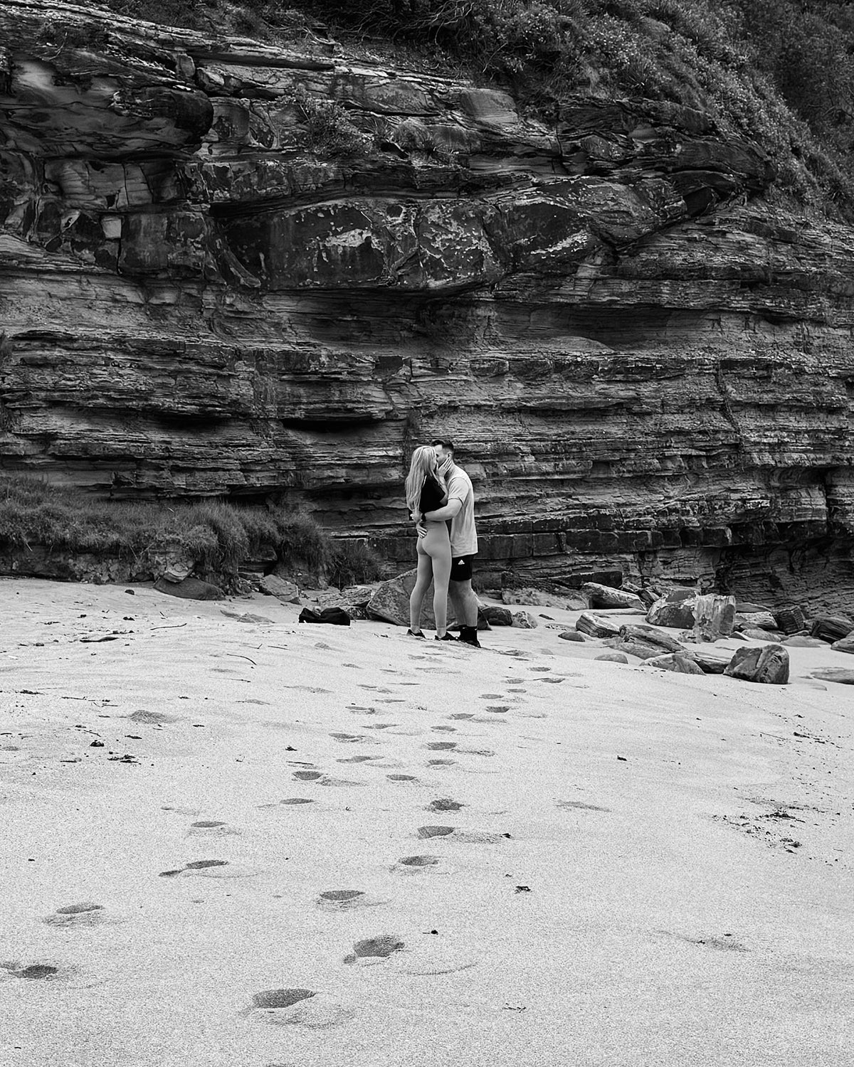 A couple embraces on a sandy beach with rocky cliffs in the background, leaving footprints behind. Black and white photo exuding romance.